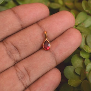 Puede incluir: Un pequeño colgante en forma de lágrima con una piedra preciosa de color rojo intenso facetada, engastada en un marco de metal dorado pulido. El colgante incluye una sencilla anilla dorada en la parte superior para sujetarlo a una cadena.