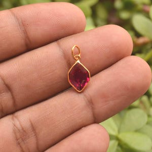 Puede incluir: Un pequeño colgante en forma de cometa con una piedra preciosa de color rojo intenso facetada, engastada en un marco de metal dorado pulido. El colgante incluye una pequeña anilla circular dorada en la parte superior para su sujeción.