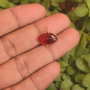 May include: A gold pendant shaped like a pair of lips featuring a smooth, translucent deep red gemstone set within a thin gold frame. The pendant includes a small gold bail at the top for attachment and a tiny decorative gold bead at the bottom centre.