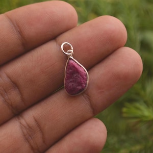 May include: A teardrop-shaped pendant featuring a raw, deep pinkish-red gemstone set in a simple silver-toned metal bezel. The pendant includes a small circular silver-toned jump ring at the top for attachment to a chain or cord.