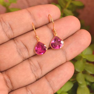 Peut inclure: Une paire de boucles d'oreilles pendantes dorées avec des pierres précieuses roses vibrantes en forme de poire à facettes. Chaque boucle d'oreille est suspendue à un crochet doré simple, mettant en valeur une finition claire et polie.
