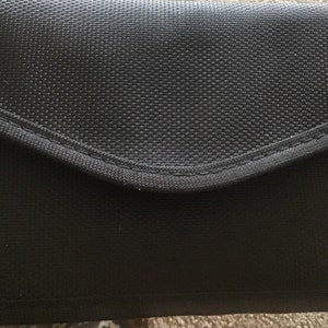 May include: A black fabric pouch with a flap closure. The pouch has a smooth, textured surface and a subtle sheen.