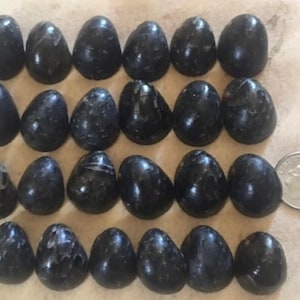 May include: A collection of twenty-one polished, dark gray, egg-shaped stones. The stones vary slightly in size and have a smooth, reflective surface. A U.S. dime is included for size comparison. The stones are arranged in a grid pattern.