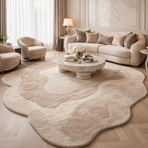 May include: An irregularly shaped area rug featuring a layered, wavy design in varying shades of beige and cream. The rug has a soft, textured pile and is positioned on a light-colored wooden floor in a room with a sofa, chairs, and a coffee table.