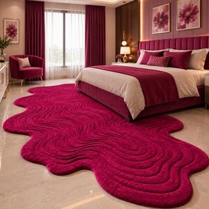 May include: A large, deep magenta area rug with an irregular, organic, wavy shape and a textured, rippled surface pattern. The rug features a thick, plush pile and covers a significant portion of the floor in front of a bed in a bedroom setting.
