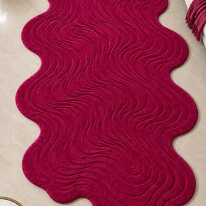 May include: A deep magenta area rug with an irregular, organic, wavy shape. The surface features a textured, rippled pattern of concentric, flowing lines that mimic the appearance of liquid movement across the entire length of the rug.
