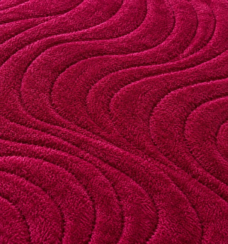 May include: A deep magenta rug featuring a textured, high-pile surface with a repeating pattern of raised, undulating wave lines that create a rhythmic, flowing visual effect across the entire fabric surface of the floor covering.