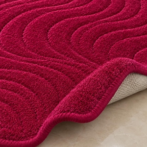 May include: A deep red bath mat featuring a textured, wavy, embossed pattern across its surface. The mat has a thick, plush pile and a finished, rounded edge. A corner of the mat is folded back to reveal a light-colored, woven fabric backing.
