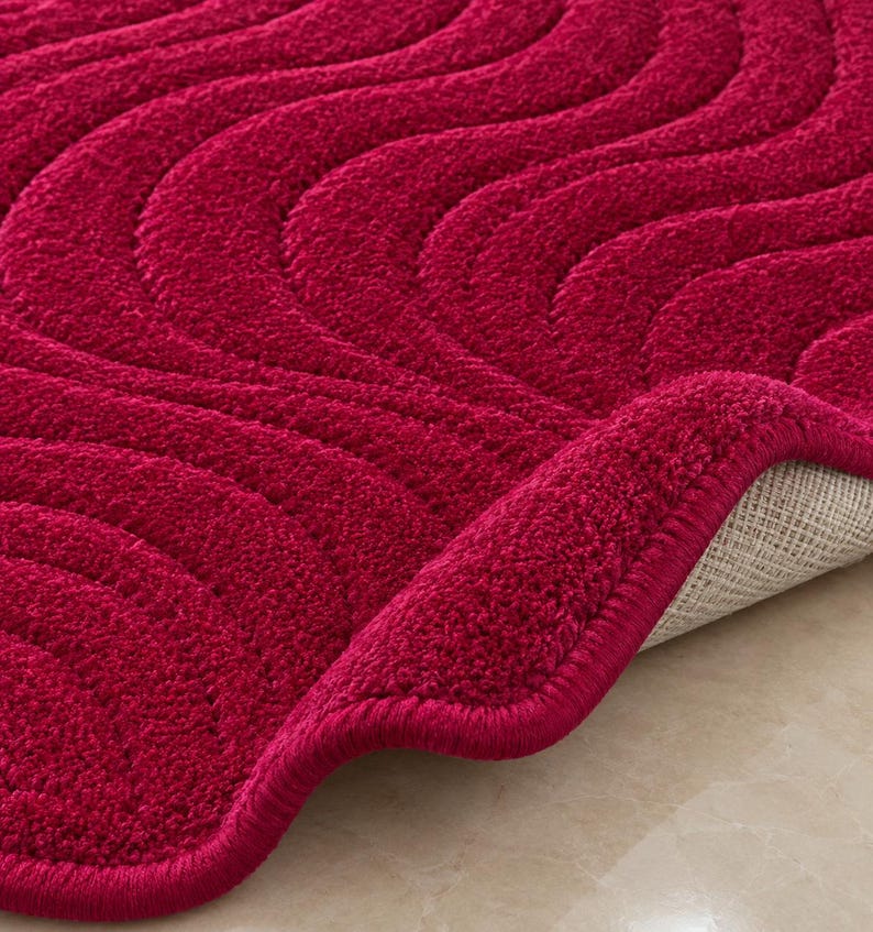 May include: A deep red bath mat featuring a textured, wavy, embossed pattern across its surface. The mat has a thick, plush pile and a finished, rounded edge. A corner of the mat is folded back to reveal a light-colored, woven fabric backing.