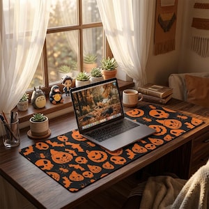 May include: A rectangular desk mat featuring a repeating pattern of orange jack-o'-lanterns, skulls, bats, and spiders on a black background. A silver laptop is centered on the mat, which is placed on a dark wood desk near a window.