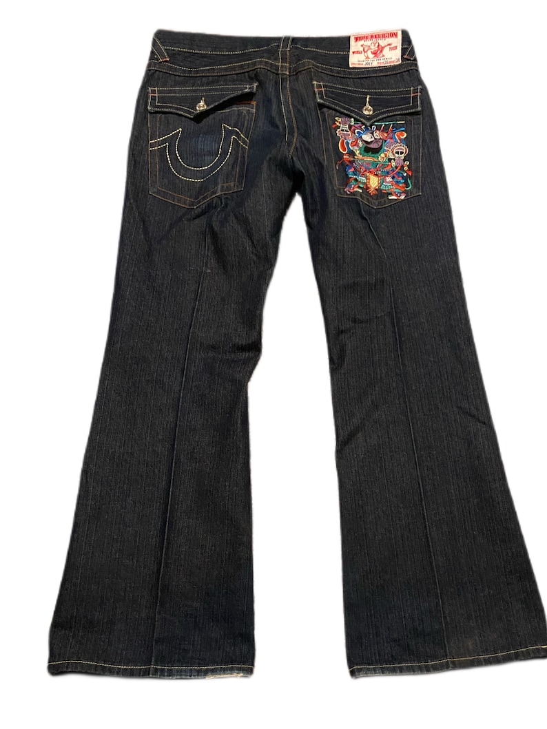 May include: Dark blue denim jeans with a large embroidered patch on the back right pocket. The patch features a colorful design with skulls, flowers, and other details. The jeans have a bootcut style.