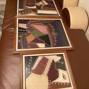May include: Three framed patchwork quilt squares with a variety of colors and textures, including brown, green, purple, and white. The frames are made of wood and have a dark brown finish.