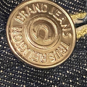 May include: A gold-colored metal button with the text "True Religion Brand Jeans" around the edge and a stylized "U" in the center.