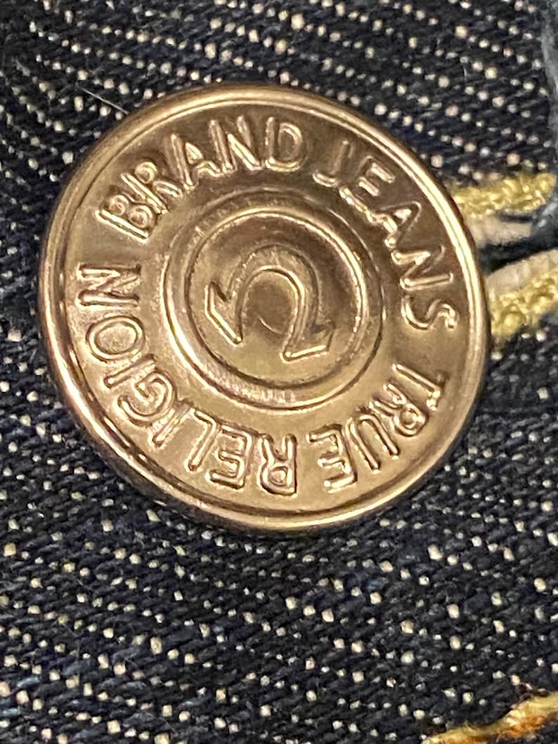 May include: A gold-colored metal button with the text "True Religion Brand Jeans" around the edge and a stylized "U" in the center.