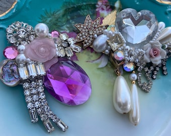Puede incluir: Dos broches vintage con una mezcla de pedrería, perlas y vidrio. Un broche presenta una rosa rosa y un gran cristal morado, mientras que el otro tiene un cristal en forma de corazón y una rosa rosa más pequeña.
