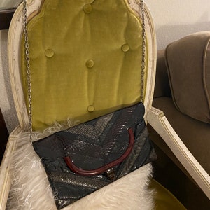 Puede incluir: Un clutch negro y plateado de piel de serpiente con un asa superior marrón. El clutch tiene una correa de cadena y está sentado sobre una manta de piel blanca.