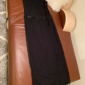 May include: A black, floor-length, sleeveless dress with a beaded waistband. The dress is made of a sheer fabric and is hanging on a brown leather sofa.