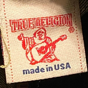 May include: A tan fabric label with red text that reads "TRUE RELIGION" and a red logo of a person with a guitar. The label also has blue text that reads "made in USA".