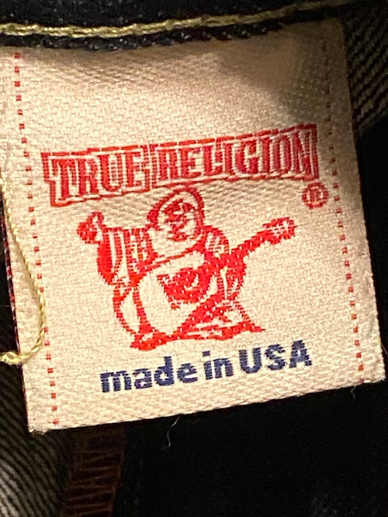 May include: A tan fabric label with red text that reads "TRUE RELIGION" and a red logo of a person with a guitar. The label also has blue text that reads "made in USA".