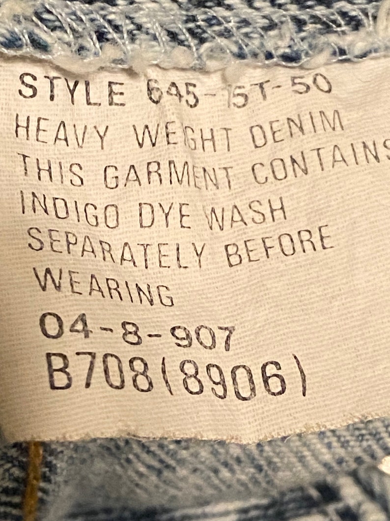 K&ouml;nnte beinhalten: Ein wei&szlig;es Etikett mit schwarzem Text, der "STYLE 645-1ST-50 HEAVY WEIGHT DENIM THIS GARMENT CONTAINS INDIGO DYE WASH SEPARATELY BEFORE WEARING 04-8-907 B708(8906)" lautet.