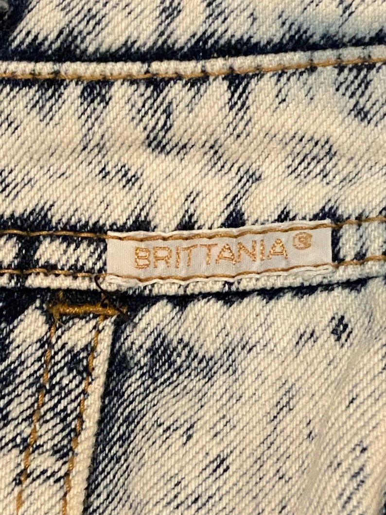 K&ouml;nnte beinhalten: Nahaufnahme eines wei&szlig;en und blauen Jeansstoffes mit einem goldenen Etikett, auf dem "BRITANNIA&reg;" steht.