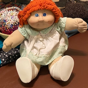 May include: A Cabbage Patch Kid doll with a red knitted hat and a light blue and white dress. The doll has blue eyes and a rosy complexion.
