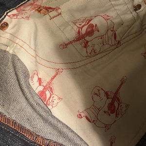 May include: Close-up of the inside of a pair of dark blue denim jeans. The lining is white with a repeated pattern of a red cartoon figure playing a guitar.