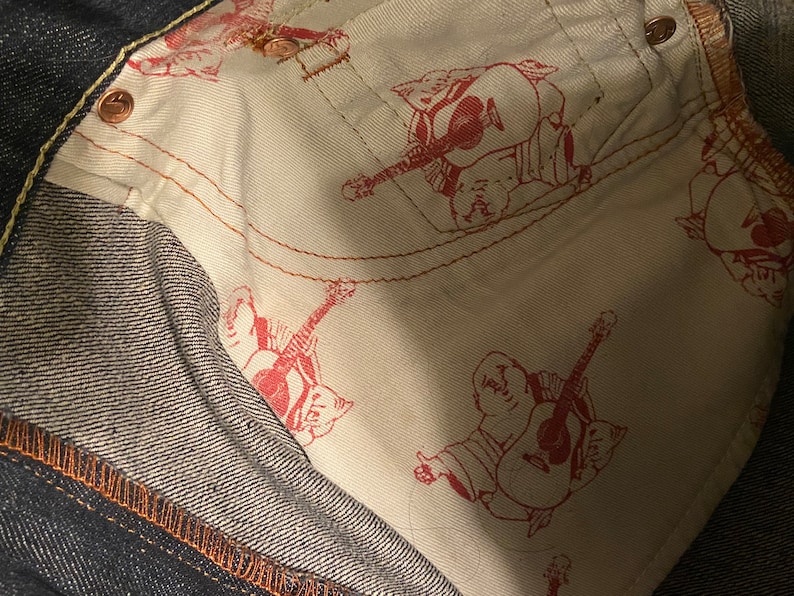 May include: Close-up of the inside of a pair of dark blue denim jeans. The lining is white with a repeated pattern of a red cartoon figure playing a guitar.