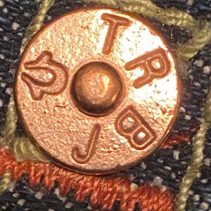 May include: Close-up of a copper-colored button with the letters "TRB" and "J" embossed on it.