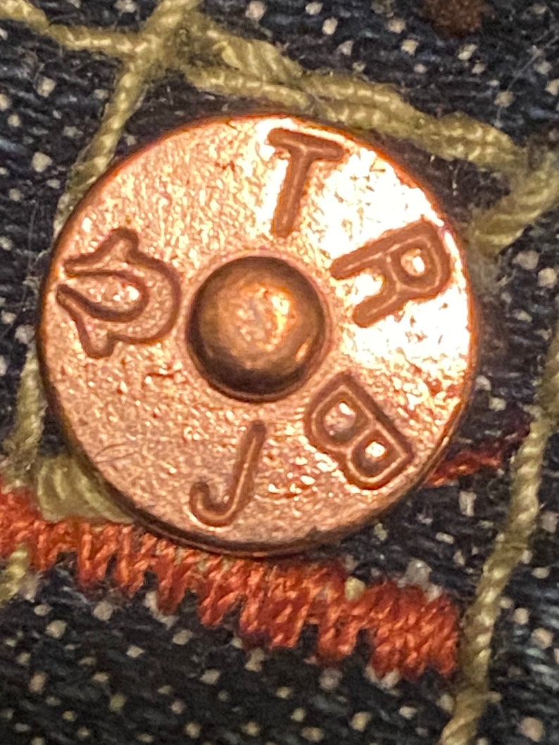 May include: Close-up of a copper-colored button with the letters "TRB" and "J" embossed on it.