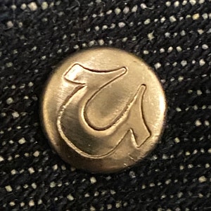 May include: A close-up of a gold-colored metal button with the letter "U" embossed on it.