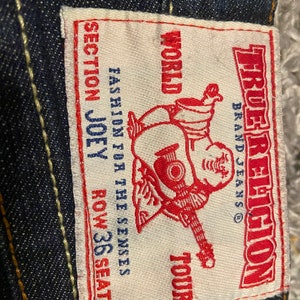 May include: A white label with red text that says "True Religion" and "World Tour" with a cartoon character holding a guitar. The label also says "Fashion for the Senses" and "Section Joey Row 36 Seat 34".