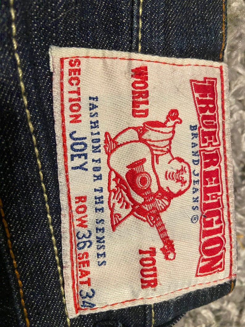 May include: A white label with red text that says "True Religion" and "World Tour" with a cartoon character holding a guitar. The label also says "Fashion for the Senses" and "Section Joey Row 36 Seat 34".