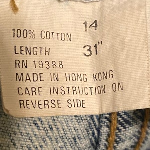 K&ouml;nnte beinhalten: Ein wei&szlig;es Etikett mit schwarzem Text, der "100% Baumwolle", "L&auml;nge", "RN 19388", "14", "31"", "Hergestellt in Hongkong" und "Pflegehinweise auf der R&uuml;ckseite" lautet.