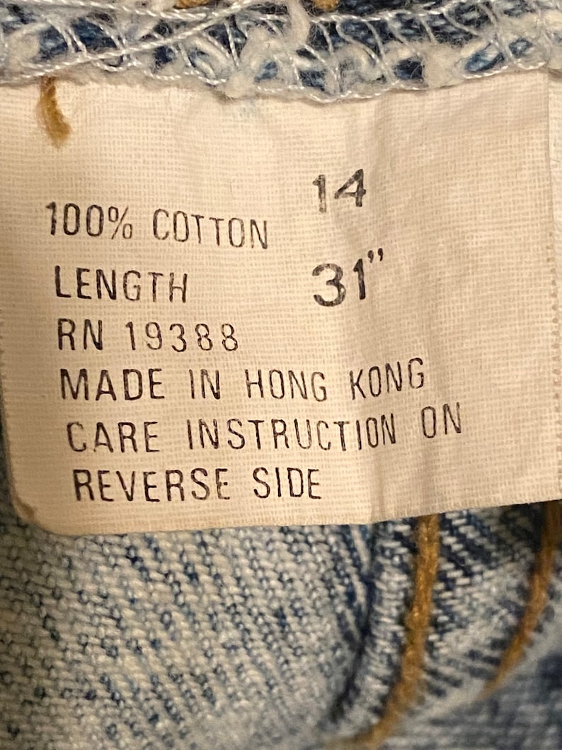 K&ouml;nnte beinhalten: Ein wei&szlig;es Etikett mit schwarzem Text, der "100% Baumwolle", "L&auml;nge", "RN 19388", "14", "31"", "Hergestellt in Hongkong" und "Pflegehinweise auf der R&uuml;ckseite" lautet.