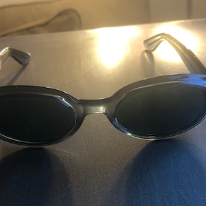May include: A pair of oval-shaped sunglasses with dark lenses and a clear gray frame.