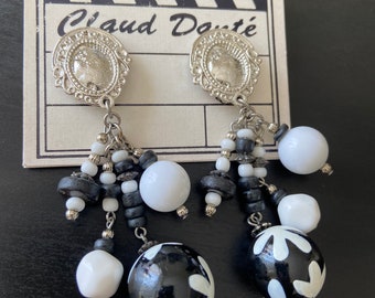 May include: A pair of dangle earrings with black and white beads, a silver charm, and a black and white patterned ball at the bottom. The earrings are on a card that says "Claude Doutré".
