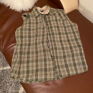 May include: A sleeveless plaid shirt in shades of green, brown, and white. The shirt has a button-up front and a single pocket on the left chest.