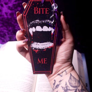 May include: A coffin-shaped bookmark with a black background, red border, and the text BITE ME in red. The center features a graphic of white lips with fangs and red blood drips. A purple tassel with a clear bead and a small silver charm is attached.