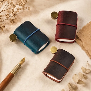 Op de afbeelding: Drie kleine in leer gebonden notitieboekjes in blauw, donkerrood en bruin, elk vastgezet met een gekleurde elastische band en voorzien van een rond messingkleurig metalen accent aan de zijkant. Een houten vulpen met een gouden penpunt ligt ernaast.