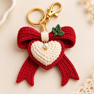 May include: A crochet keyring featuring a large red bow and a smaller cream-coloured heart centred in the middle. A single white pearl bead sits below the centre knot of the bow, and a small green crocheted leaf is attached to the top right. A gold metal ring and lobster clasp are attached to the top.