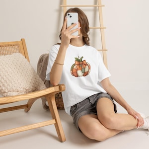Puede incluir: Una camiseta blanca de manga corta con un gráfico central de una calabaza estilo patchwork. El diseño de la calabaza incorpora varios patrones en tonos naranja, marrón y crema, incluyendo cuadros y lunares, rematado con un pequeño lazo de cuadros naranja.