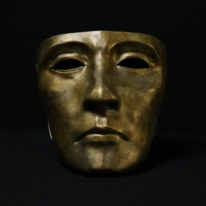 Peut inclure: Un masque facial de couleur bronze présentant une expression neutre avec des arcades sourcilières marquées, des ouvertures oculaires en amande, un nez droit et des lèvres fermées. La surface a une finition métallique marbrée.