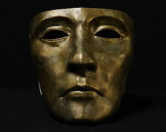 Könnte beinhalten: Eine bronzefarbene Gesichtsmaske mit neutralem Ausdruck, ausgeprägten Brauenbögen, mandelförmigen Augenöffnungen, einer geraden Nase und geschlossenen Lippen. Die Oberfläche weist ein fleckiges, metallisches Finish mit Lichtschattierungen auf.