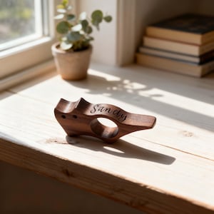 Puede incluir: Un servilletero de madera de color marrón oscuro con una forma suave y contorneada y una abertura circular en el centro. La palabra Sandy está grabada en letra cursiva en la superficie superior de la madera, que muestra un veteado natural.