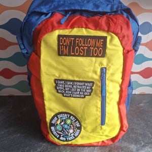 Può includere: Uno zaino blu, rosso e giallo con una toppa superiore che recita STAY WEIRD, una toppa rettangolare con la scritta DON'T FOLLOW ME I'M LOST TOO, una toppa testuale più lunga e una toppa circolare con WHAT DOESN'T KILL YOU MUTATES AND TRIES AGAIN.