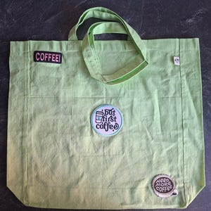 May include: A light green tote bag featuring three embroidered patches. One rectangular patch reads COFFEE!, a circular patch displays a coffee cup with the text but first coffee, and a third circular patch reads need more coffee.