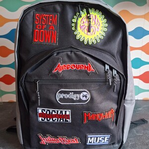 May include: A black backpack featuring various band patches including System of a Down, Blind Melon, Airbourne, The Prodigy, Social Distortion, Manowar, Judas Priest, and Muse. The bag has a large main compartment and a smaller front zippered pocket.