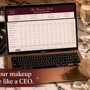 May include: A laptop displaying The Glamour Desk budget planner dashboard with financial data, surrounded by makeup brushes, an eyeshadow palette, a glass of champagne, and a white peony. The text reads: Run your makeup empire like a CEO.