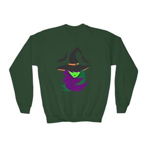 May include: A dark forest green crewneck sweatshirt featuring a central graphic of a witch with bright green skin, purple hair, and a black pointed hat with an orange band, accompanied by two small black bat silhouettes flying above the hat.